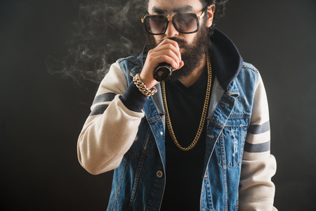 Young man with beard and sunglasses vaping an electronic cigarette. Vaper hipster smoke vaporizerの写真素材