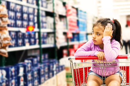 Sale, consumerism and people concept - happy little girl pensive in shopping cart at supermarketの写真素材