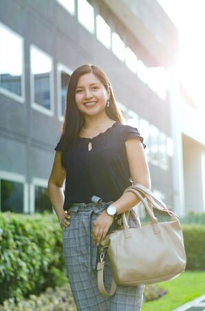 Businesswoman summer suit for office. The fashionable woman wears high heels, plaid cotton pants and a beige outdoor bag. Buildings backgroundの写真素材