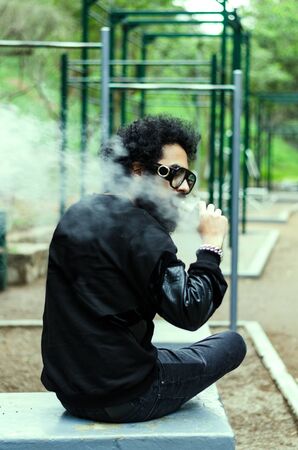 Man with a beard smokes an electronic cigarette sit on a bench in the park
