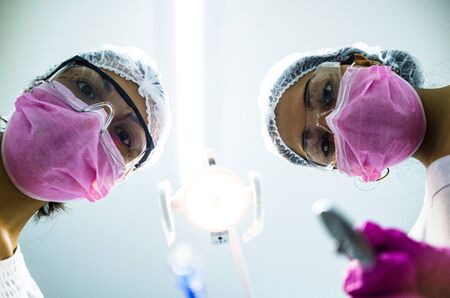 Bottom view of two women dentists in surgical mask holding tools and looking at camera. Patient...
