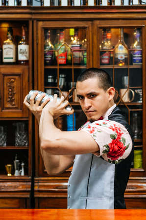 Young handsome smiling barman in bar interior shaking and mixing alcohol cocktail. Professional...
