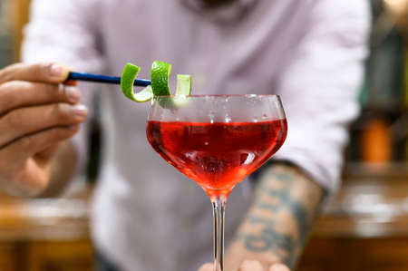close-up of glass with red drink that barman accurate decorate with orange peel.