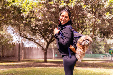 A charming young woman walks in nature, with a backpack on her shoulder, from which her dog peeks...