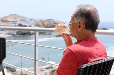 Mature man sitting on a balcony overlooking the sea with a cocktail.
