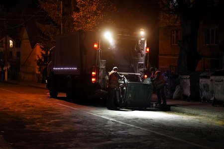 Garbage collecting truck is parked on city street. Driver prepares garbage container for loading into truck. Dark time of day.の写真素材