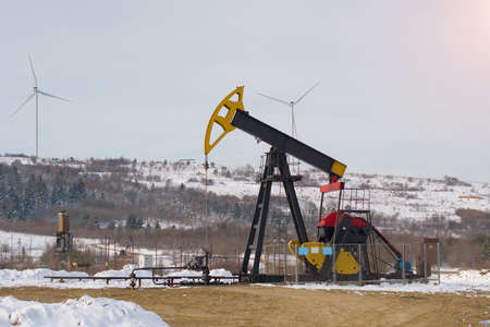 Oil pump. Oil and gas industry equipment. Oil field pump jack and oil refinery in the winter with snow, wind turbine mountains and forest in backgroundの写真素材