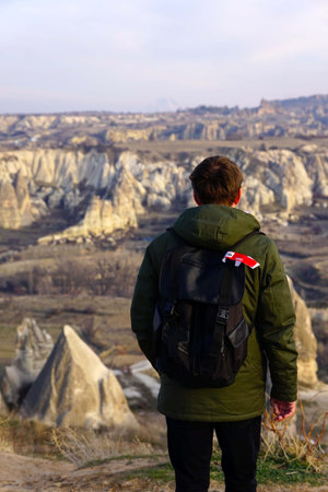 Young Male Tourist Hiking with Backpacks on Top of Rocky Canyon Valley in Front of a city Goreme Cappadocia view in the Mountains of National Park.の写真素材