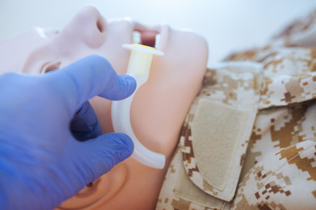Blue-gloved hand measuring an oropharyngeal airway tube against a medical training mannequin in a camouflage uniform to check the correct size for airway management.の写真素材