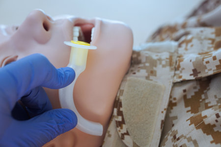 Blue-gloved hand measuring an oropharyngeal airway tube against a medical training mannequin in a camouflage uniform to check the correct size for airway management.の写真素材
