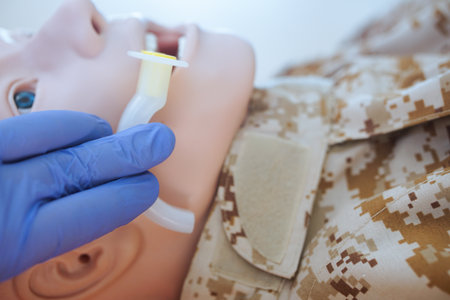 Blue-gloved hand measuring an oropharyngeal airway tube against a medical training mannequin in a camouflage uniform to check the correct size for airway management.の写真素材