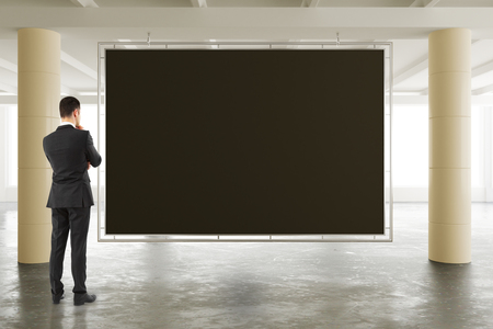 Businessman looking at blank blackboard in sunny spacious hangar area with concrete floor, mock upの写真素材
