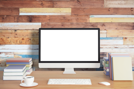 Blank computer screen with keyboard, piles of books and cup of coffee on wooden table and wooden wall, mock upの写真素材