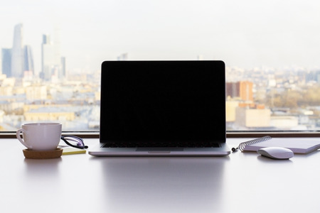 Front view of blank laptop screen, coffee cup and other items on white desktop with sunlit city in the background. Mock upの写真素材