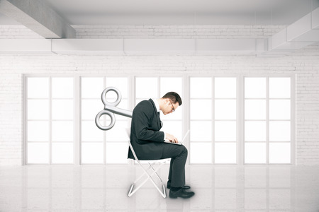 Businessman with a wind-up key on his back sitting on chair and using laptop in white brick room. Concept of controlの写真素材