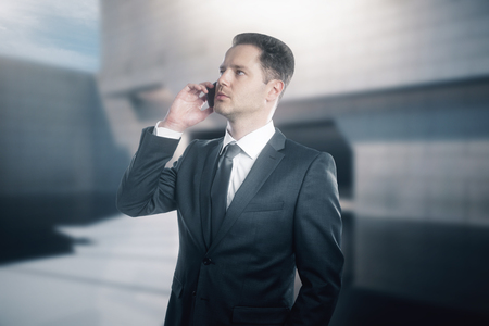 Attractive caucasian businessman talking on phone. Blurry concrete tile exterior in the backgroundの写真素材