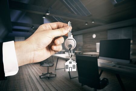 Realtor hand holding key with house keychain on office interior background. Real estate and mortgage conceptの写真素材