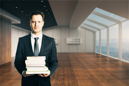 Handsome businessman holding stack of books in unfurnished modern interior. Education concept. 3D Renderingの写真素材