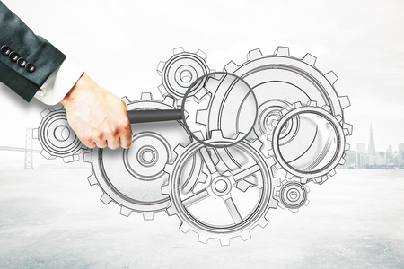 Businessman hand holding magnifier over cogwheel sketch on abstract city background. Research conceptの写真素材