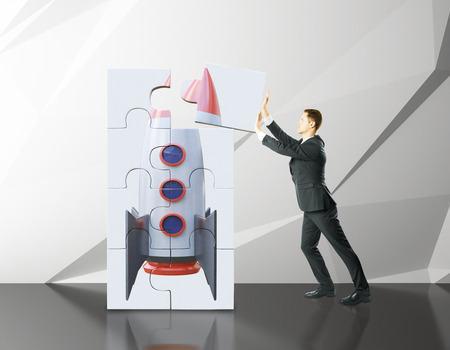 Young businessman adding last piece to rocket ship puzzle in concrete interior. Project start up and development process. 3D Renderingの写真素材