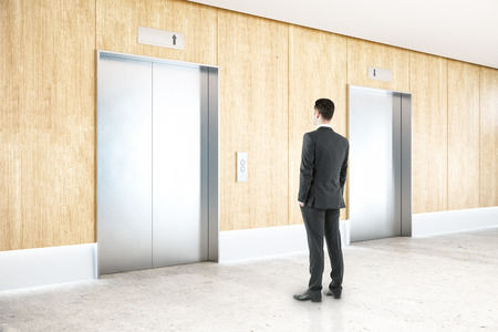 Thoughtful businessman in interior with two silver elevators, wooden wall, concrete floor and...