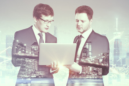 Two handsome european businessmen using laptop together on city background. Technology concept. Double exposureの写真素材