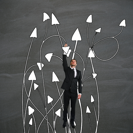 Abstract image of young businessman hanging on tangled arrows. Chalkboard background. Different direction conceptの写真素材