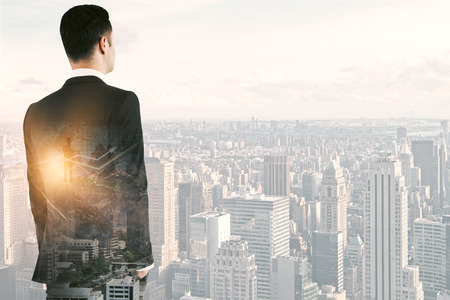 Back view of thoughtful young businessman with forex chart on bright city background with copy space and sunlight. Finance concept. Double exposureの写真素材