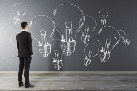 Back view of thoughtful young businessman in interior looking at concrete wall with drawn lamps.. Idea and creativity conceptの写真素材