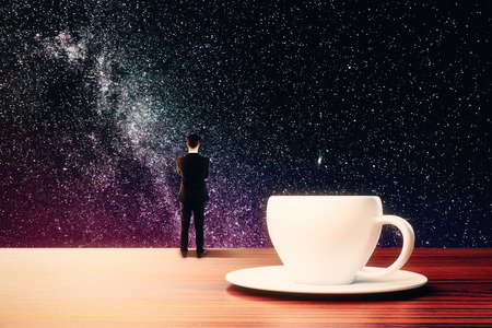 Back view of abstract thoughtful tiny businessman standing on wooden desktop with coffee cup on saucer and looking at space night sky. Creativity concept. 3D Renderingの写真素材
