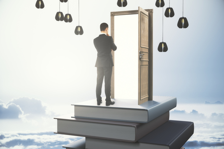 Back view of thoughtful young businessman standing on abstract book tower with open door on sky background. Career concept. 3D Renderingの写真素材