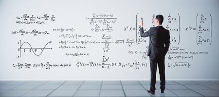 Back view of young businessman writing mathematical formulas on concrete wall in interior. Science conceptの写真素材