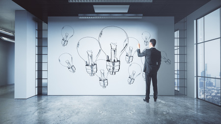 Back view of young businessman drawing lamps on concrete wall of grunge office interior with city view. Innovation and idea concept. 3D Renderingの写真素材