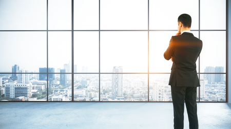 Back view of thoughtful young businessman standing in modern concrete office with panoramic city view and daylight. Research concept. 3D Rendering の写真素材