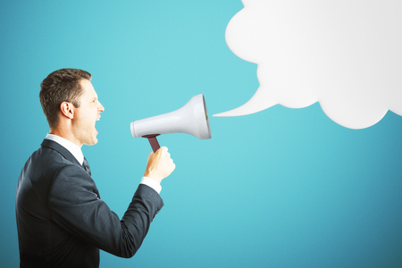 Side view of handsome young businessman screaming into megaphone on blue background with empty speech/thought cloud. Mock up. Communication and message conceptの写真素材