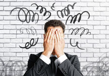Stressed businessman covering face on white brick wall background with scribble. Stress concept 