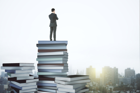 Back view of young businessman standing on abstract book pile and looking into the distance on blurry city background. Education and research concept. 3D Rendering の写真素材