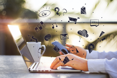 Side view of young businessman using laptop and smartphone with abstract social media sketch on blurry outdoor background. Communication, technology and network concept の写真素材