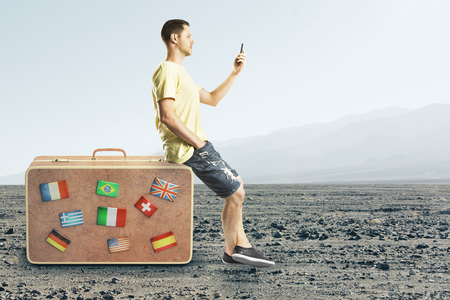 Side view of handsome european guy with smartphone sitting on suitcase on beautiful landscape background. Travel and social media conceptの写真素材