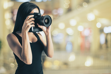 Pretty young european girl taking photograph with professional camera on blurry lights background. Hobby and occupation conceptの写真素材