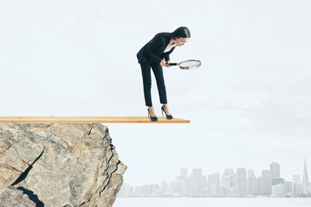 Businesswoman with magnifier looking down from cliff. City background. Research and risk conceptの写真素材