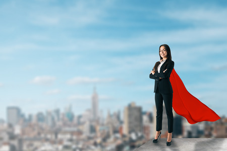 Beautiful young businesswoman with red hero cape on rooftop with blurry city view and daylight. Confidence and power conceptの写真素材