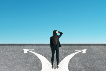 Back view of young businesswoman standing on road with arrows. Sky background. Different direction conceptの写真素材
