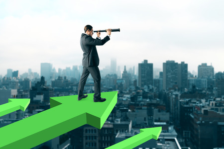 Businessman standing on abstract red arrows and using telescope to look into the distance on city background. Growth and research conceptの写真素材