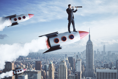 Businessman using telescope while standing on launching rocket. City background. Research and startup conceptの写真素材