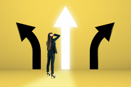 Young businesswoman with arrow choice on yellow background. Different direction and leadership conceptの写真素材