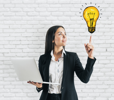Attractive smiling young european woman holding laptop and abstract drawn lamp on brick wall background. Communication and innovation conceptの写真素材
