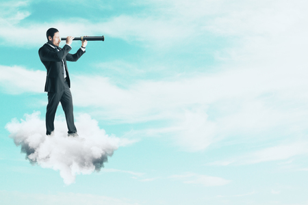 Side view of young businessman on cloud using telescope to look into the distance on bright blue sky background. Vision and research conceptの写真素材