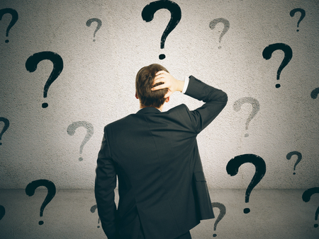 Back view of young businessman on concrete wall background with question marks. Confusion and doubt conceptの写真素材