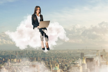 Attractive young european businesswoman using laptop while sitting on cloud on sky city background. Freelance conceptの写真素材